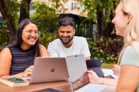 The University of Queensland’s Master of Writing, Editing and Publishing. Three UQ students from a range of cultural backgrounds sit at an outdoor table discussing their work, with laptops, books and papers beside them.