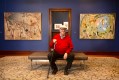 Old man in red jumper and cane in gallery setting with paintings. William Robinson