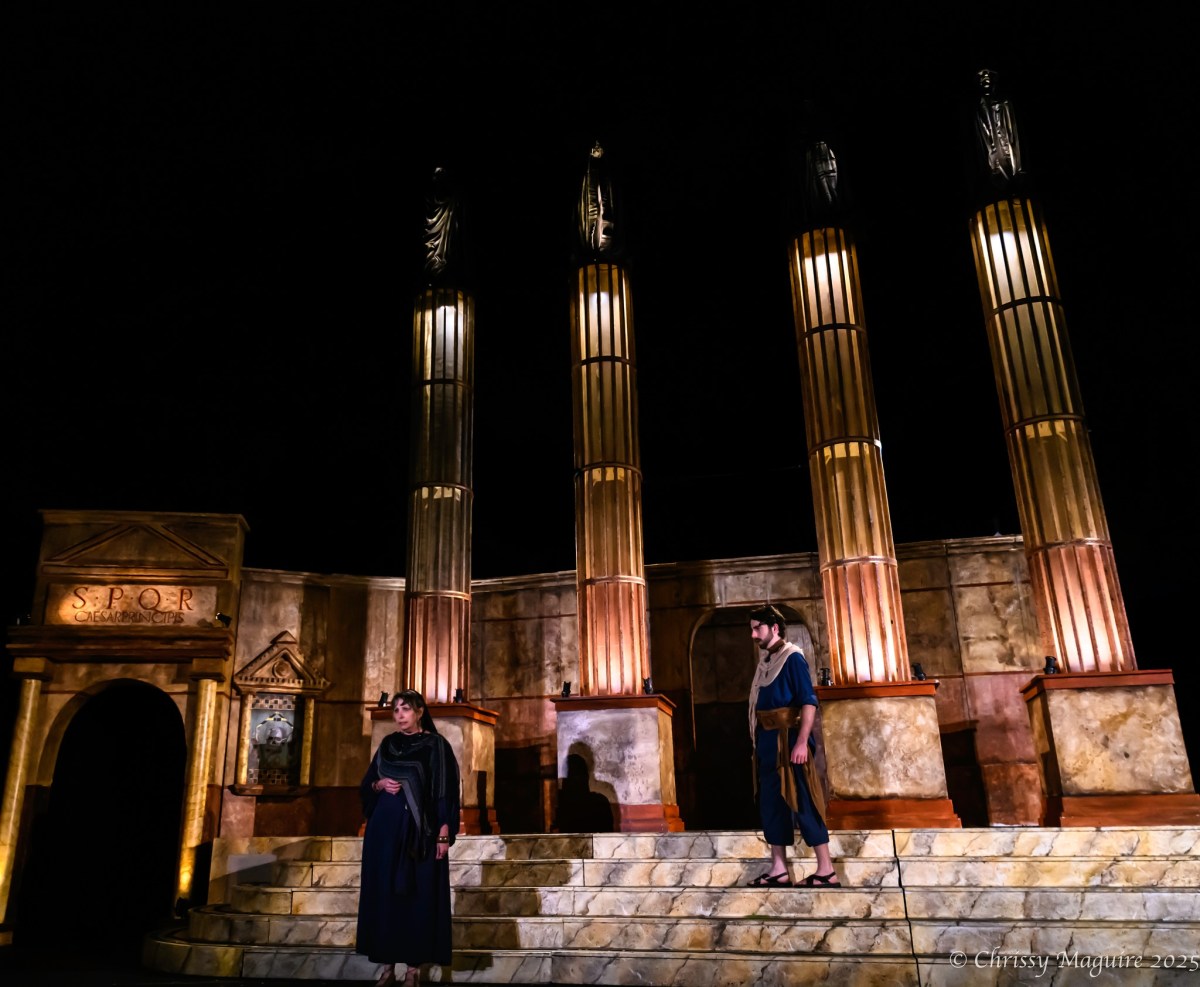 Two people in black standing on steps. There are big columns behind them in a production of Caesar.