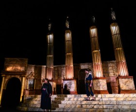 Two people in black standing on steps. There are big columns behind them in a production of Caesar.