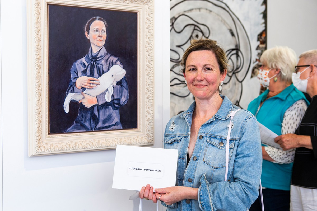 A middle-aged Caucasian woman standing next to her portrait inside a gallery. More opportunities, grants and awards.