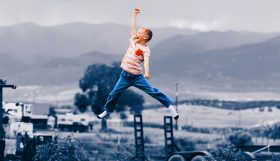 A red-headed young boy leaps high into the air, one fist raised triumphantly above his head. He wears blue jeans, white socks and no shoes, and an orange and white striped t-shirt. The photo's background, a semi-rural landscape, is rendered in shades of blue, against which the boy stands out dramatically. The photo illustrates ArtsHub's weekly On the move column, a round-up of Australian arts sector appointments.