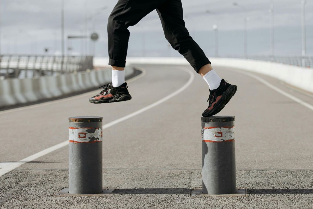 A close-up photograph of a person jumping from bollard to bollard on a stretch of road; the photo shows only their legs and feet. The person is wearing black pants, white socks and black and red sneakers. The photo illustrates Artshub's weekly On the Move column, which summarises the week's arts sector appointments across Australia.