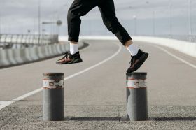 A close-up photograph of a person jumping from bollard to bollard on a stretch of road; the photo shows only their legs and feet. The person is wearing black pants, white socks and black and red sneakers. The photo illustrates Artshub's weekly On the Move column, which summarises the week's arts sector appointments across Australia.