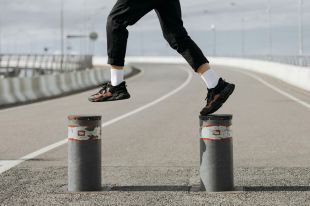 A close-up photograph of a person jumping from bollard to bollard on a stretch of road; the photo shows only their legs and feet. The person is wearing black pants, white socks and black and red sneakers. The photo illustrates Artshub's weekly On the Move column, which summarises the week's arts sector appointments across Australia.