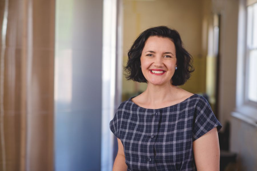 Polyglot Theatre's Kath Fyffe, a dark haired, fair-skinned woman in a short-sleeved, blue and grey checked dress, smiles at the camera.