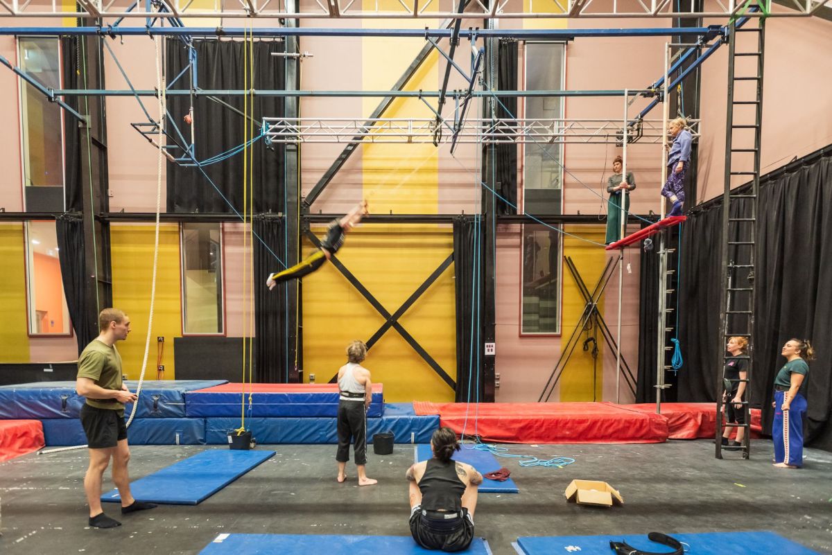 Circus artists training in The Hub at Circus Centre Melbourne. A trapeze artist swings, their photo blurred by motion, as other artists watch on.