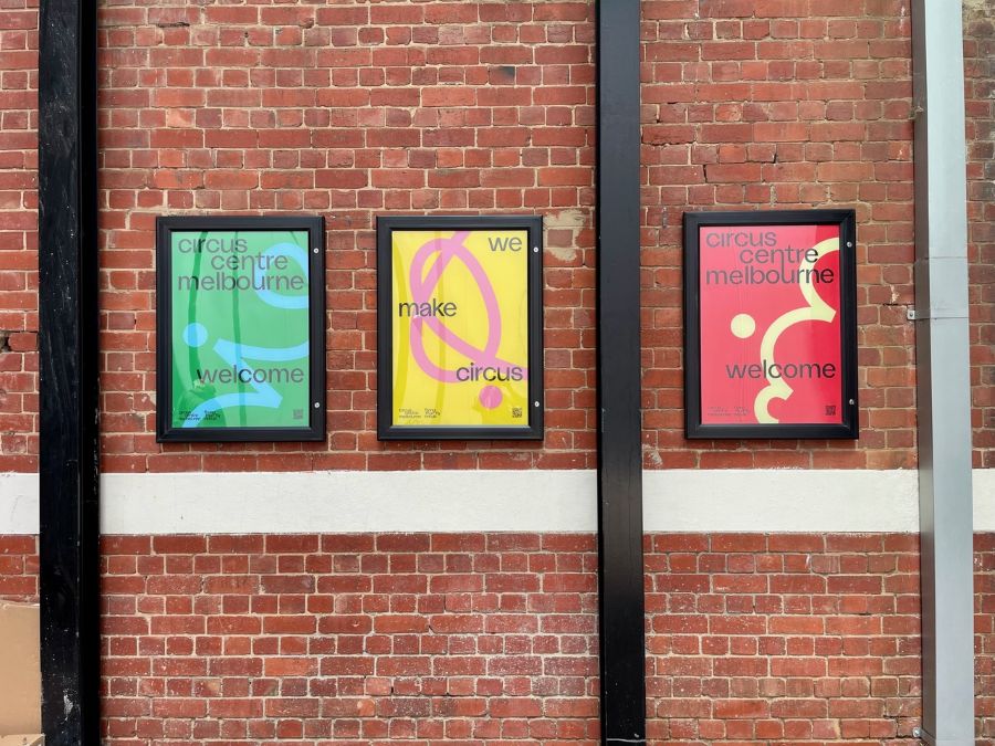 Circus Centre Melbourne branding at the building's northern entrance. The photograph depicts three colourful signs hanging on a red brick wall. from left to right they read: 'circus centre melbourne welcome', 'we make circus' and 'circus centre melbourne welcome'.