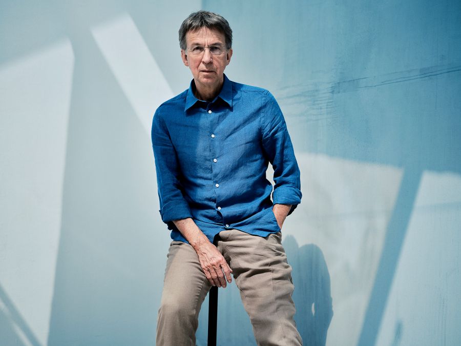 Chief Conductor and Artistic Director of the Tasmanian Symphony Orchestra, Eivind Aadland. An older, fair-skinned man with greying hair and wearing frameless glasses, a blue shirt and tan-coloured slacks. He sits with his left hand in his pocket and his right hand resting across his right thigh.