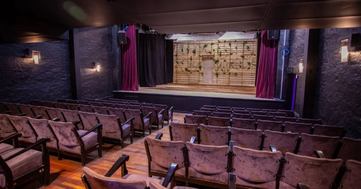 Ad Astra's 150-seat proscenium arch Galaxy Theatre. The photograph shows several rows of padded chairs on a raked floor facing a stage; its red curtains are open showing the stage floor and a simple set representing a lattice screen garden.