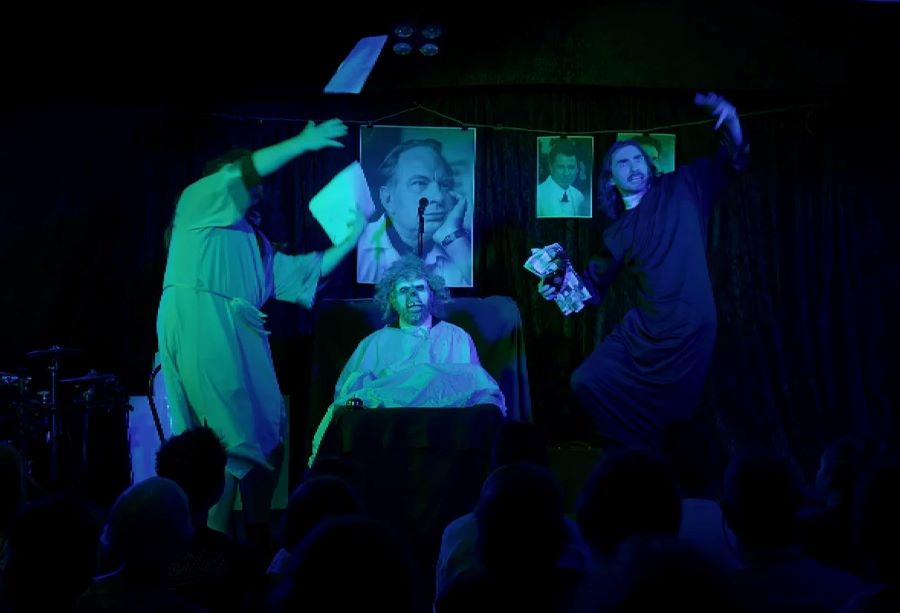 A scene from 'George Glass' Scientology the Musical' at Melbourne Fringe. A demonically-masked figure lies in a hospital bed under a blue-green light while two robed figures cavort comically around it.