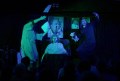 George Glass' 'Scientology the Musical' at Melbourne Fringe 2025. A moody photograph lit in shades of green and blue showing a faux-exorcism underway. Robed and gowned figures stand either side of a bed containing a demonically-masked person; photographs hang in the background.