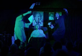 George Glass' 'Scientology the Musical' at Melbourne Fringe 2025. A moody photograph lit in shades of green and blue showing a faux-exorcism underway. Robed and gowned figures stand either side of a bed containing a demonically-masked person; photographs hang in the background.