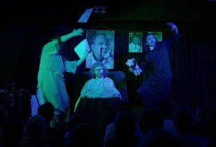George Glass' 'Scientology the Musical' at Melbourne Fringe 2025. A moody photograph lit in shades of green and blue showing a faux-exorcism underway. Robed and gowned figures stand either side of a bed containing a demonically-masked person; photographs hang in the background.