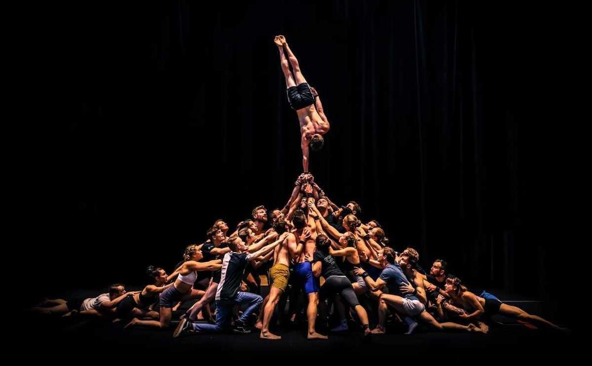 A large mass of circus performers forming an amorphous pyramid shape with their bodies on stage against a black background. The lone performer at the top of the human pyramid is balancing in a one handed handstand.
