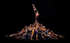 A large mass of circus performers forming an amorphous pyramid shape with their bodies on stage against a black background. The lone performer at the top of the human pyramid is balancing in a one handed handstand.