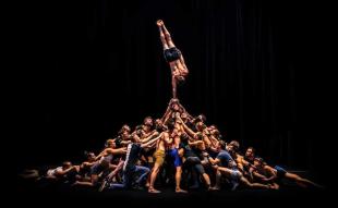 A large mass of circus performers forming an amorphous pyramid shape with their bodies on stage against a black background. The lone performer at the top of the human pyramid is balancing in a one handed handstand.