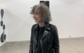 A side profile photo of Anna Schwartz (a Caucasian woman with curly gray hair) wearing a black biker’s jacket in an exhibition space with white walls.