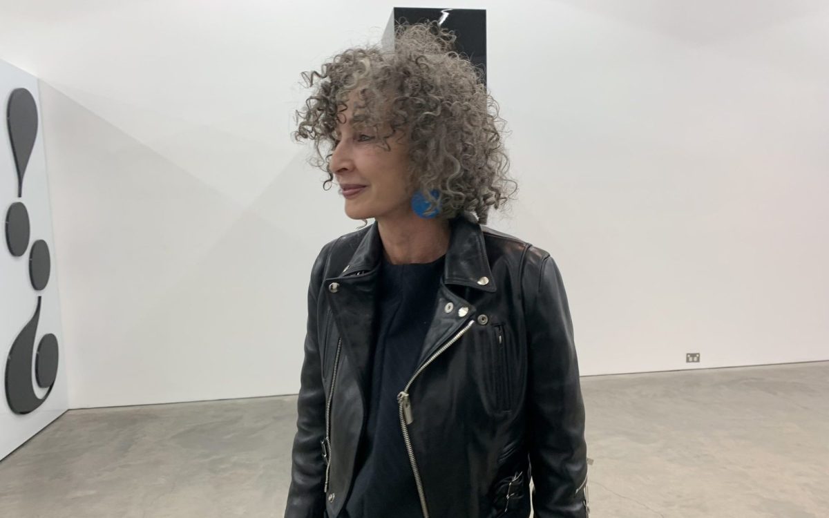 A side profile photo of Anna Schwartz (a Caucasian woman with curly gray hair) wearing a black biker’s jacket in an exhibition space with white walls.