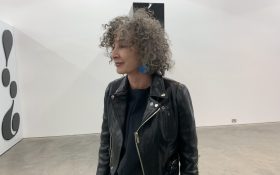 A side profile photo of Anna Schwartz (a Caucasian woman with curly gray hair) wearing a black biker’s jacket in an exhibition space with white walls.