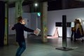 A dimly lit gallery exhibition space. To the left is a man mimicking the shape of a cross in response to an artwork of the same shape on the right. Opportunities and more.