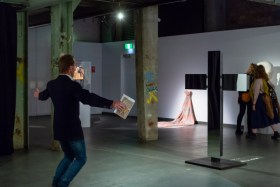 A dimly lit gallery exhibition space. To the left is a man mimicking the shape of a cross in response to an artwork of the same shape on the right. Opportunities and more.