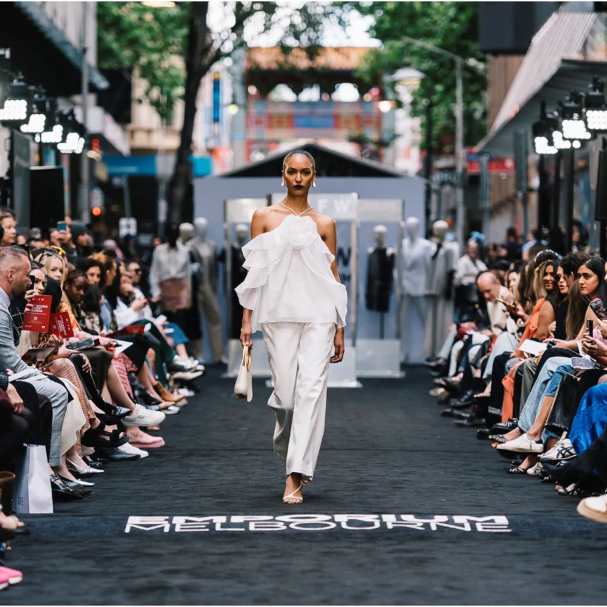 A model in white walking down an outdoor fashion runway with audiences on both sides.