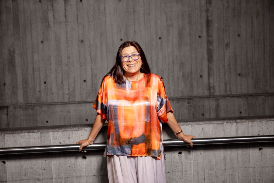 Joanne_Kee, the new announced Artistic Director of the Seymour Centre. Photo: Stefanie Zingsheim, University of Sydney. An Asian-Australian woman with glasses, long dark hair and wearing a colourful, short sleeved top leans against a metal railing and concrete balustrade.