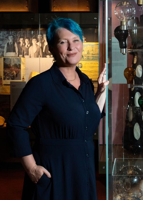 City of Newcastle's Director, Museum Archive Libraries and Learning, Julie Baird, at the Newcastle Museum. Baird is the new President of AMaGA. The photo depicts a woman in a dark blue dress with short, bright blue hair; she stands with her left hand against a glass display case and her right hand in her pocket.