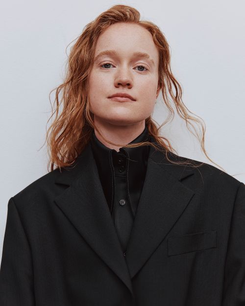 Canberra Youth Theatre alumnus Liv Hewson. The photo shows an androgynous actor with fair skin and long red hair wearing a a high-necked black shirt and jacket. They gaze cooly at the camera, their head tilted slightly to the left.