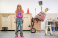 Nell, a middle-aged Caucasian woman with long curly hair and a big smile, wearing neon pink tshirt and sneakers, standing in front of some installation works in her studio. Soon to show at Heide.