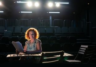 NIDA Open Studios Senior Tutor Louise Birgan. The photograph depicts a fair skinned woman with shoulder length red hair and wearing blue demim overalls over a sleeveless white and blue striped t-shirt. She is sitting on stage at a table, holding a script and is dramatically backlit; rows of theatre seats are visible in the gloom behind her.