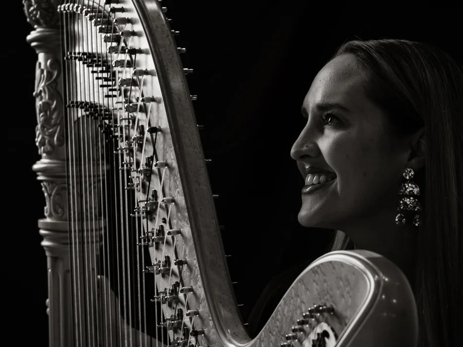 Queensland Symphony Orchestra's Principal Harp, Emily Granger. A close-up, black and white photograph showing a smiling woman with fair skin, long dark hair and wearing ornate earings, sitting in profile at a large harp.