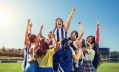 A promotional image for Queensland Theatre Company's season 2026 production, 'Strong is the new Pretty'. A group of woman photographed on an AFL oval are celebrating; a woman wearing a blue and white striped North Melbourne guensey is at the centre of the image, her left fist held aloft triumphantly.