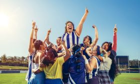 A promotional image for Queensland Theatre Company's season 2026 production, 'Strong is the new Pretty'. A group of woman photographed on an AFL oval are celebrating; a woman wearing a blue and white striped North Melbourne guensey is at the centre of the image, her left fist held aloft triumphantly.