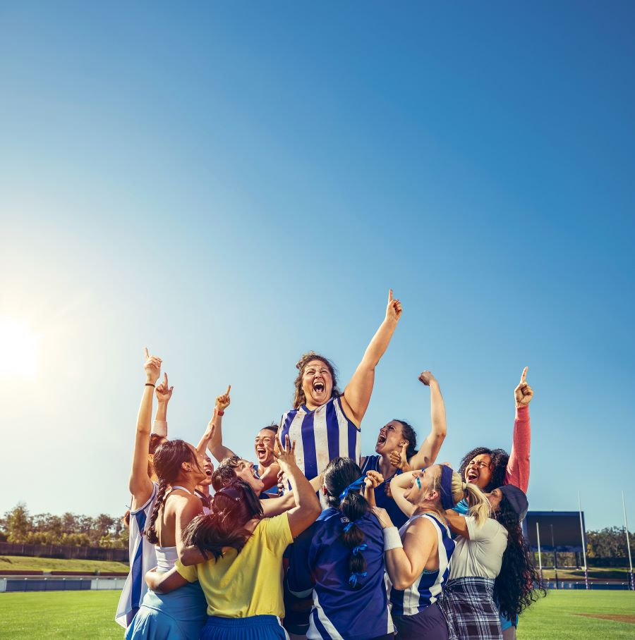 A promotional image for Queensland Theatre Company's season 2026 production, 'Strong is the new Pretty'. A group of woman photographed on an AFL oval are celebrating; a woman wearing a blue and white striped North Melbourne guensey is at the centre of the image, her left fist held aloft triumphantly.