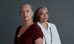 L-R: Rachael Maza and Lisa Maza in a promotional image for Malthouse Theatre's 2026 production, 'Black Light'. The photo shows two older Aboriginal woman standing beside one another but not looking at each other; they both have curly grey hair and stern, resolute expressions. The woman on the left wears a red dress; her sister standing beside her wears a white shirt and a long, beaded necklace.