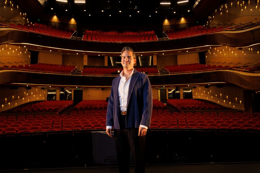 State Opera South Australia Artistic Director Dane Lam. A fair skinned man wearing a blue jacket, white collared shirt and black trousers, stands on stage smiling at the camera; an empty theatre auditorium and its seats and balconies are visible behind him.