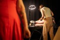 A blurry arm with tattoos in the foreground. In the background the converse clad legs of someone laying on a tattoo bed while Xani, dressed in brown, gives them a tattoo. A scene from 'Tattoo Show' by Rawcus at Melbourne Fringe 2025.