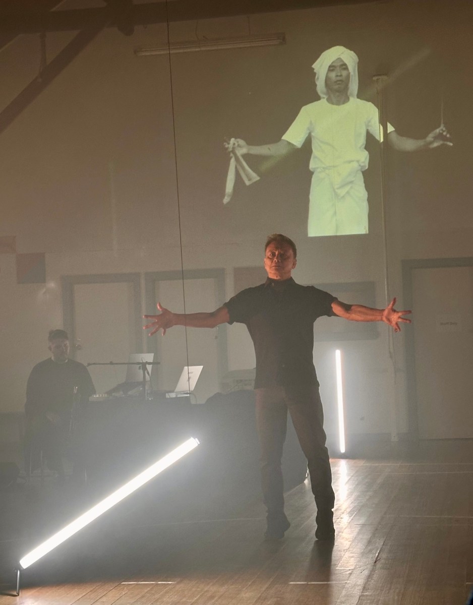 male dancer in black performing in a smokey room with projected image behind him. The Unconformity