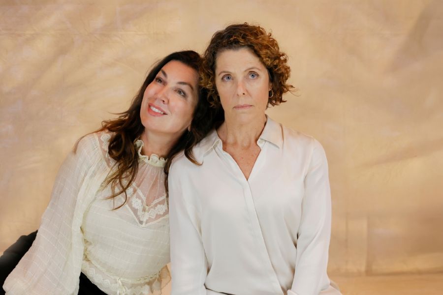 L-R: Sopranos Antoinette Halloran and Natalie Jones in a promotional image fo Victorian Opera's 2026 season world premiere producion, The Spare Room, based on Helen Garner's novel of the same name. A smiling, fair-skinned woman with long dark hair rests her head on the shoulder of a more severe-looking, fair-skinned woman with shorter, curly hair. Both women are wearing white shirts or blouses.