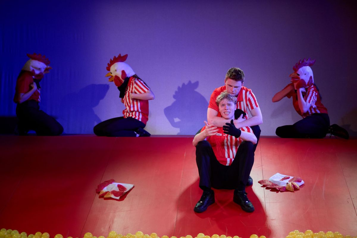 A scene from Canberra Youth Theatre's 2024 production of 'Work, But This Time Like You Mean It'. Two young men sit on a steeply raked stage, comforting one another; both wear black trousers, black shoes and red and white striped collared shirts. Behind them, three people in identical uniforms but wearing surreal chick head masks, sit at the top of the raked stage.