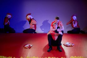 A scene from Canberra Youth Theatre's 2024 production of 'Work, But This Time Like You Mean It'. Two young men sit on a steeply raked stage, comforting one another; both wear black trousers, black shoes and red and white striped collared shirts. Behind them, three people in identical uniforms but wearing surreal chick head masks, sit at the top of the raked stage.