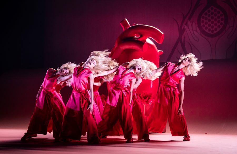 A scene from Queensland Ballet and Experience Gold Coast's 'Elastic Heart' at HOTA, 2025. Seven dancers, identically dressed in red costumes and long blonde wigs, toss their hair and sway before a model of the human heart which is larger than any of the dancers.