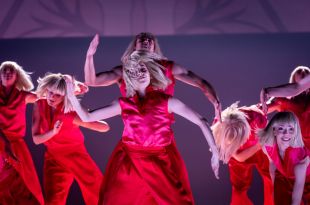 A scene from Queensland Ballet and Experience Gold Coast's 'Elastic Heart' at HOTA, 2025. Seven dancers perform, each identically dressed in red costumes and blonde wigs, recreating Sia's iconic look. Two of the central dancers stamd in the same pose, with arms extended and bent at the elbow: the right forearm and hand pointing up, the left forearm and hand pointing down.