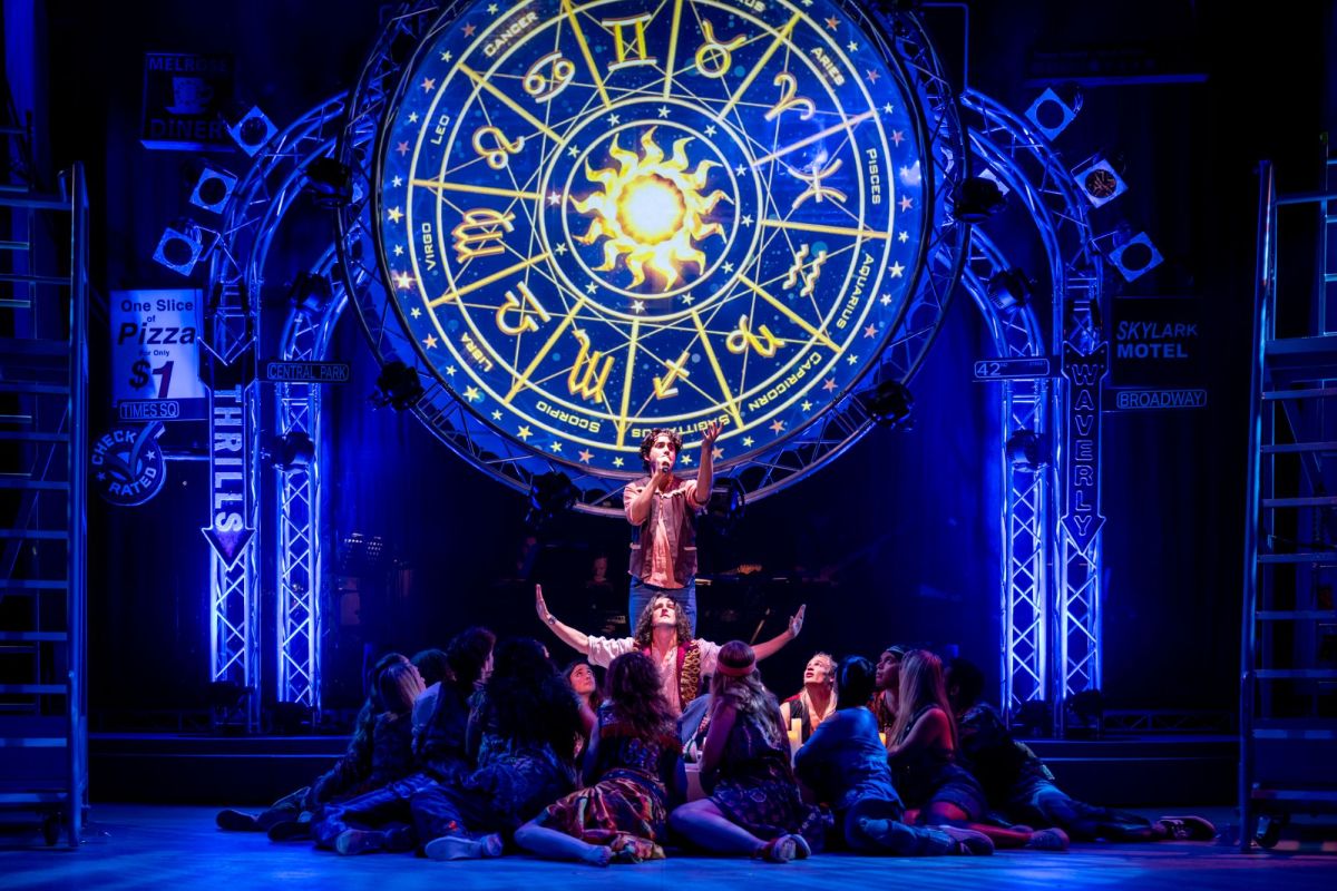 The 2025 cast of the Australian production of 'Hair'. A large, illuminated zodiac star-wheel dominates a blue-lit stage; the golden symbols of the zodiac shine brightly. Members of the cast are gathered beneath the zodiac wheel, some crouching, others with arms raised or outstretched as if gripped by religious fervour.