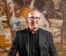 Man in black suit and glasses in formal portrait in front of painting. Jason Smith