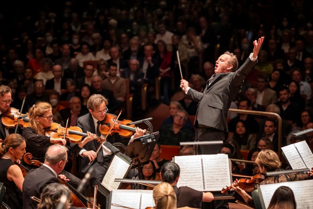 Alexander Briger conducts the Australian World Orchestra. Briger has his arms raised dramatically and his head is thrown back; orchestra members are arrayed before him, with audience members visible behind him.
