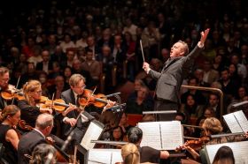 Alexander Briger conducts the Australian World Orchestra. Briger has his arms raised dramatically and his head is thrown back; orchestra members are arrayed before him, with audience members visible behind him.