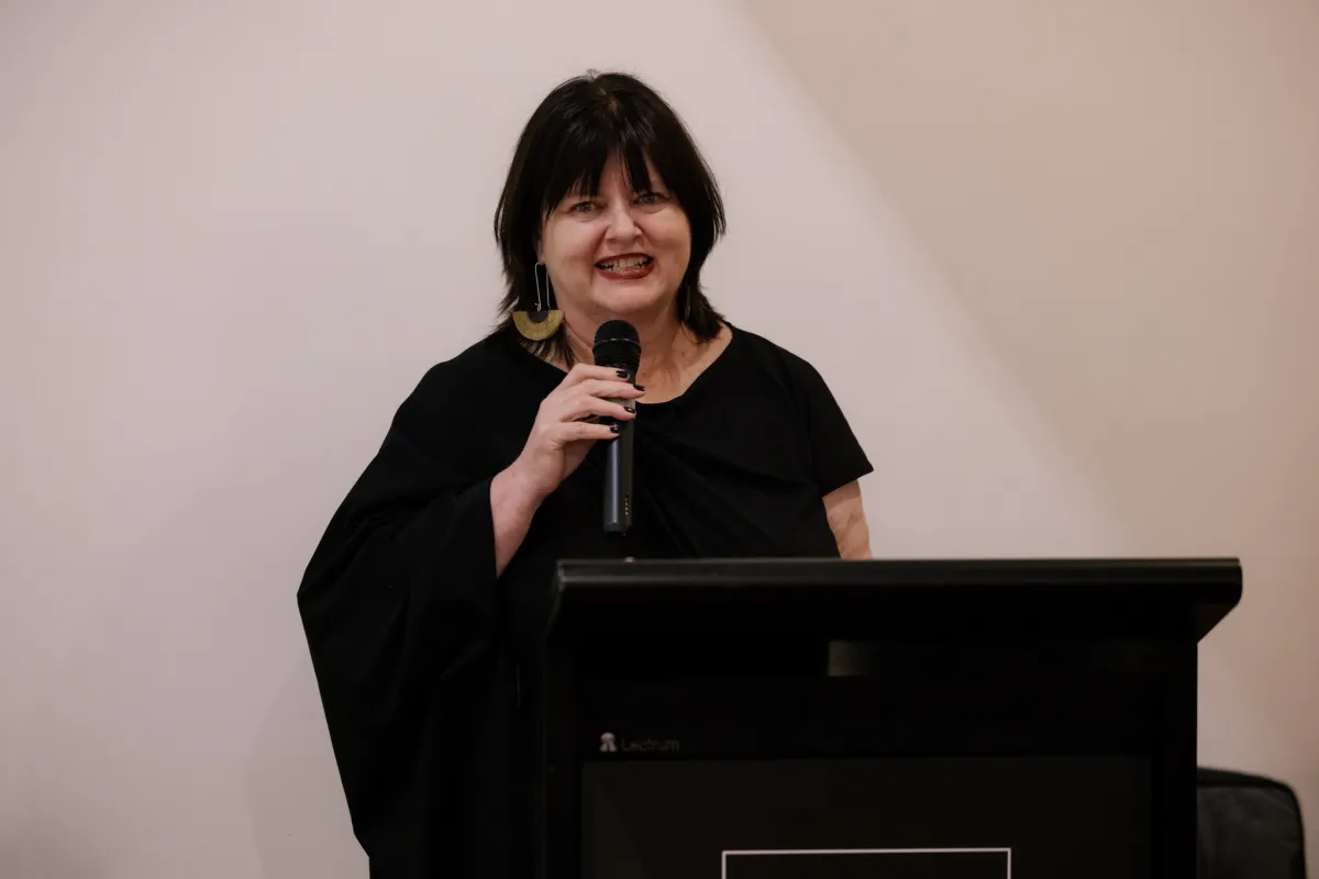woman with dark hair in black dress, smiling and giving speech in gallery. ADC